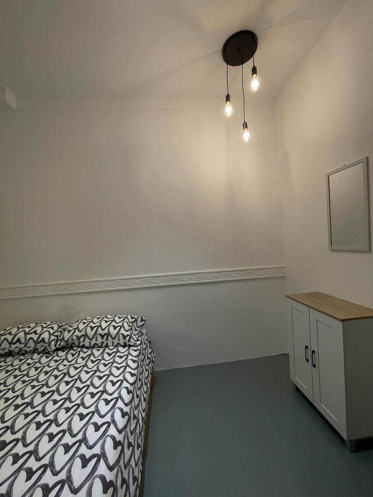A bedroom features a patterned bedspread with heart motifs over a spacious bed. A simple wooden side table with cabinet doors stands nearby. Soft lighting is provided by exposed bulbs hanging from the ceiling, illuminating the light-colored walls.