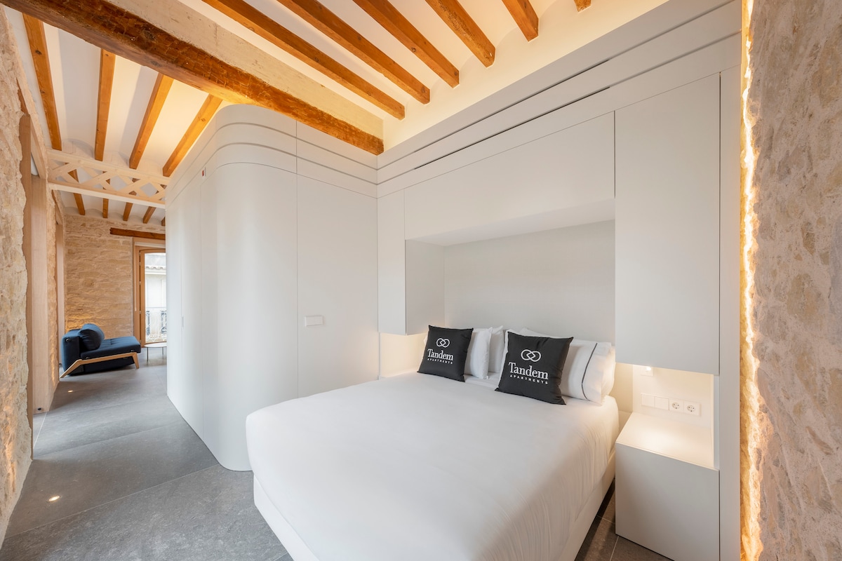 A spacious bedroom features a comfortable bed with soft linens and decorative cushions. Built-in storage offers a sleek design, while natural light fills the room through a large window. Exposed wooden beams add a touch of character to the modern aesthetic.