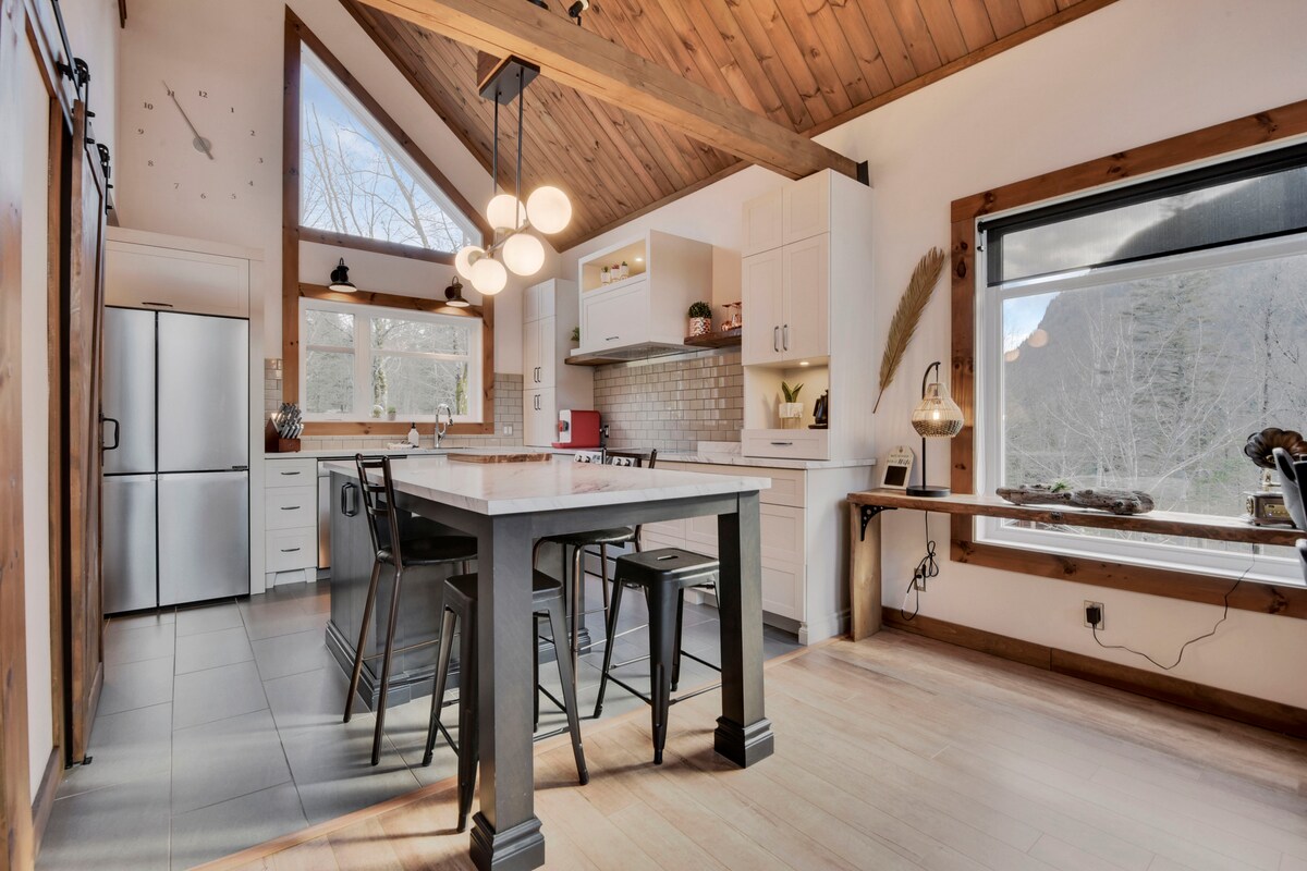 A spacious kitchen area features modern appliances, including a stainless steel refrigerator and built-in oven. A large island with seating invites social interaction. Light floods in through large windows, highlighting the natural wood accents and high, sloped ceiling, creating a warm and functional cooking space.