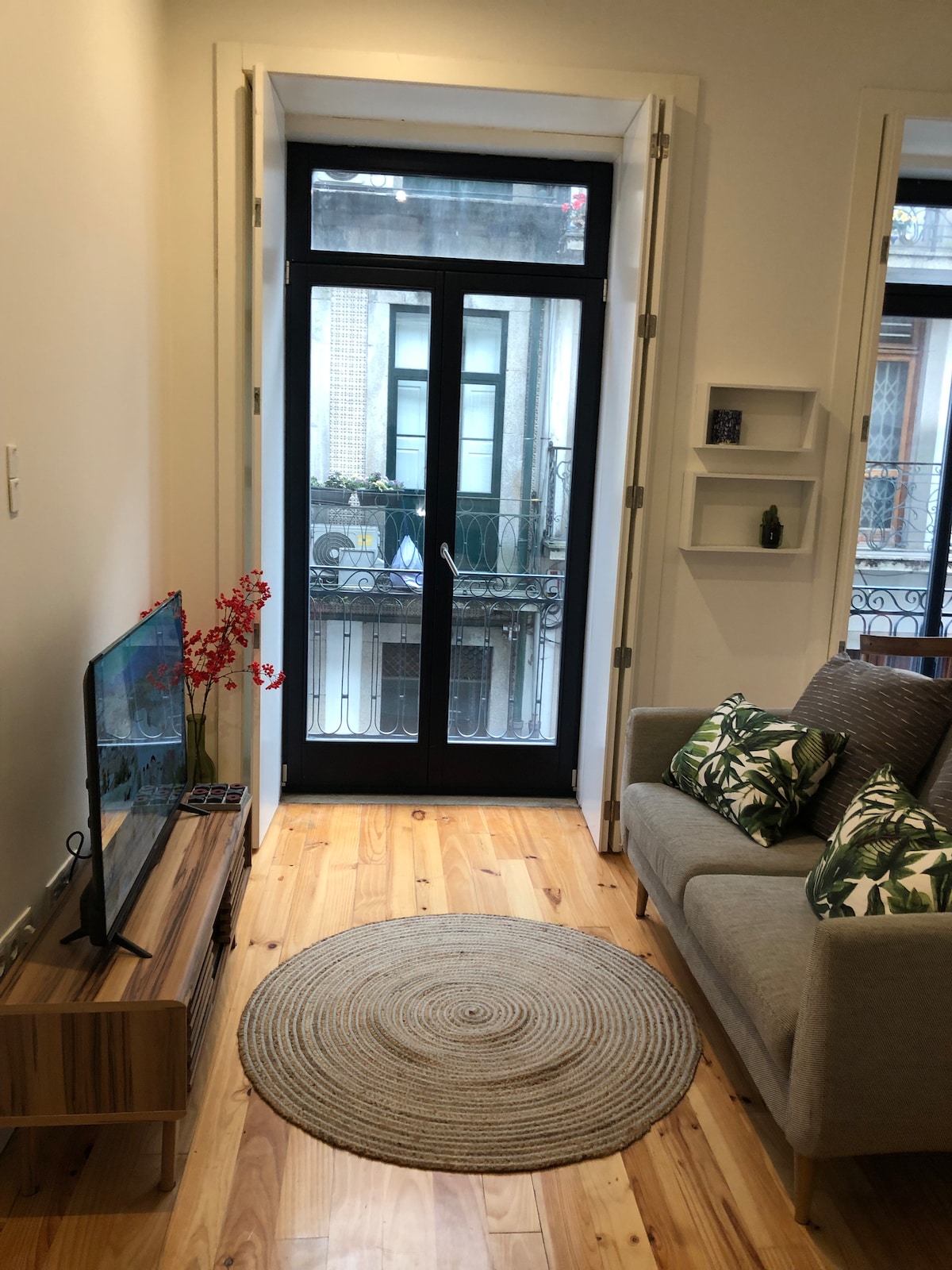 A cozy living area is highlighted by a gray sofa adorned with green leaf-patterned cushions. A small round rug sits on the wooden floor, complementing the warm tones. Large doors provide access to a balcony overlooking the street, adding natural light to the space.