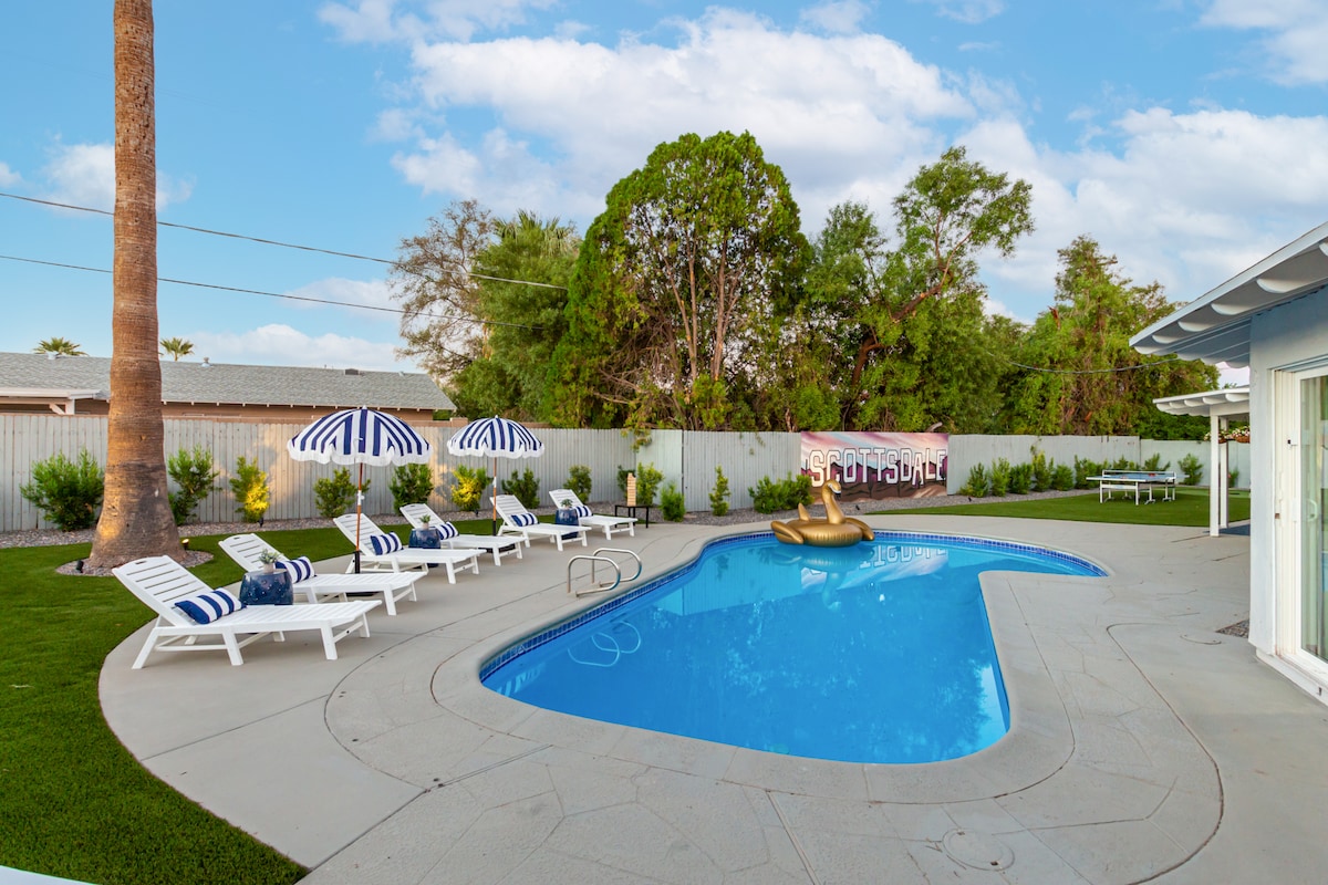 A spacious backyard features a heated pool surrounded by beach loungers with striped umbrellas. Near the pool, a mural displaying the word 'Scottsdale' adds a playful touch. Lush greenery borders the yard, creating a serene environment for relaxation and entertainment.
