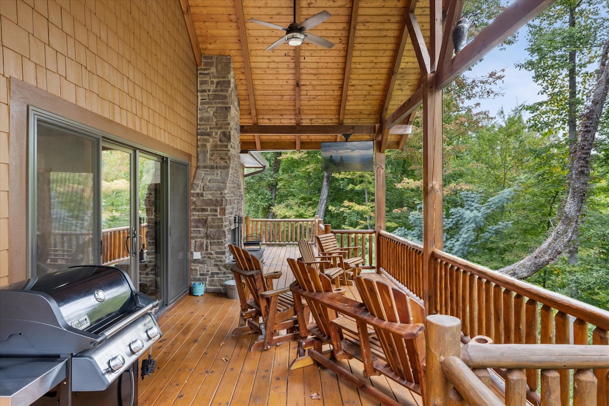 An expansive deck features a gas grill and multiple wooden rocking chairs, inviting relaxation amidst a serene wooded backdrop. Large windows reveal views of the surrounding trees, while a ceiling fan provides comfort on warm days. The structure incorporates rustic wooden elements.
