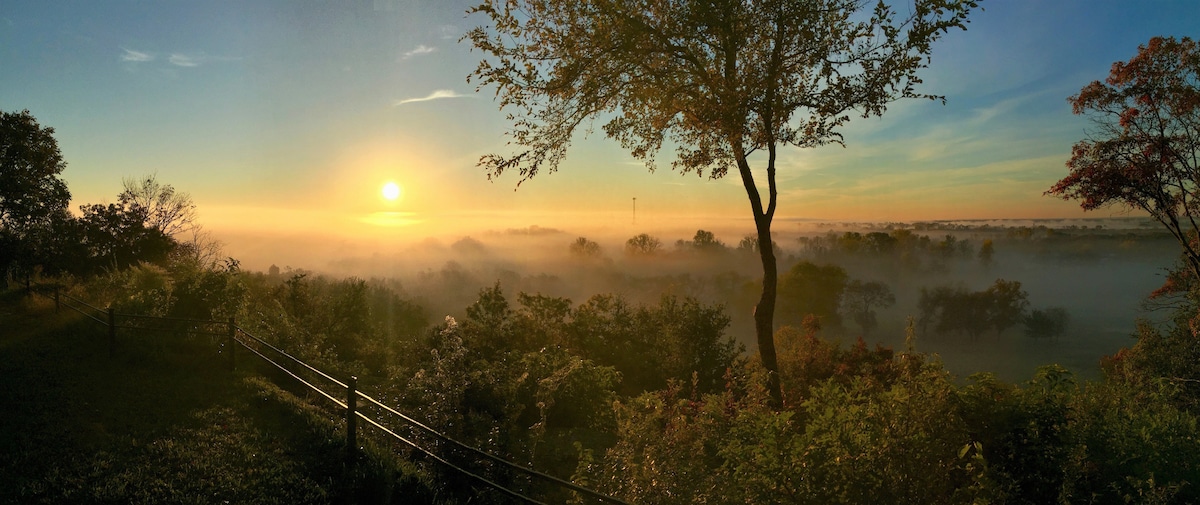 A panoramic view captures the sunrise over a misty landscape, with the sun rising above the horizon. Lush greenery is visible in the foreground, complemented by trees that outline the scene. A wooden fence delineates the area, creating a sense of tranquility and natural beauty.