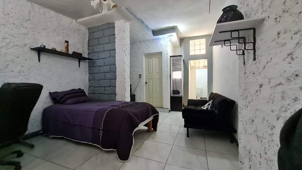 A cozy sleeping area features a spacious bed with dark purple bedding, complemented by a black sofa. Textured white walls and a gray accent wall provide a unique backdrop. A small shelf holds decorative items, and a mirror reflects natural light from a nearby window.