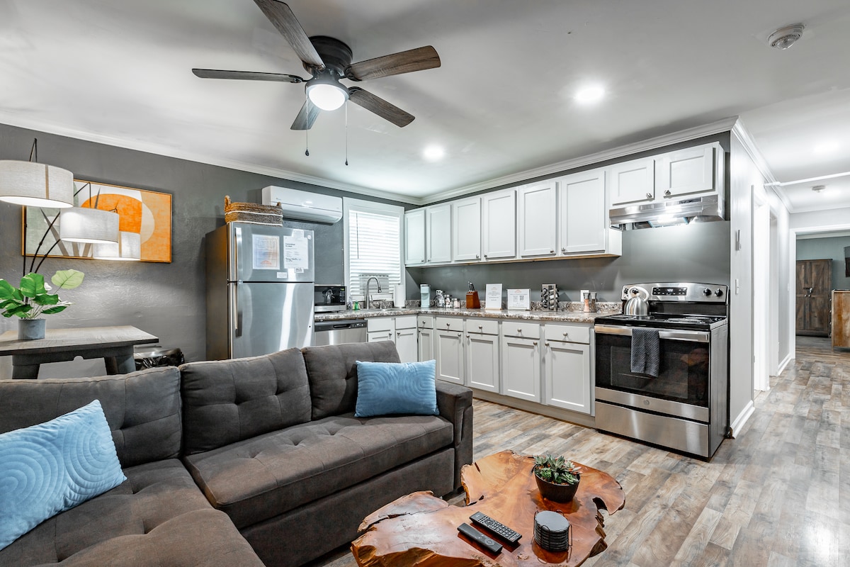 A cozy living area features a spacious gray sectional sofa with light blue cushions, and a wooden coffee table. The kitchen, equipped with stainless steel appliances and white cabinetry, is visible in the background. Soft lighting creates a welcoming ambiance throughout the room.