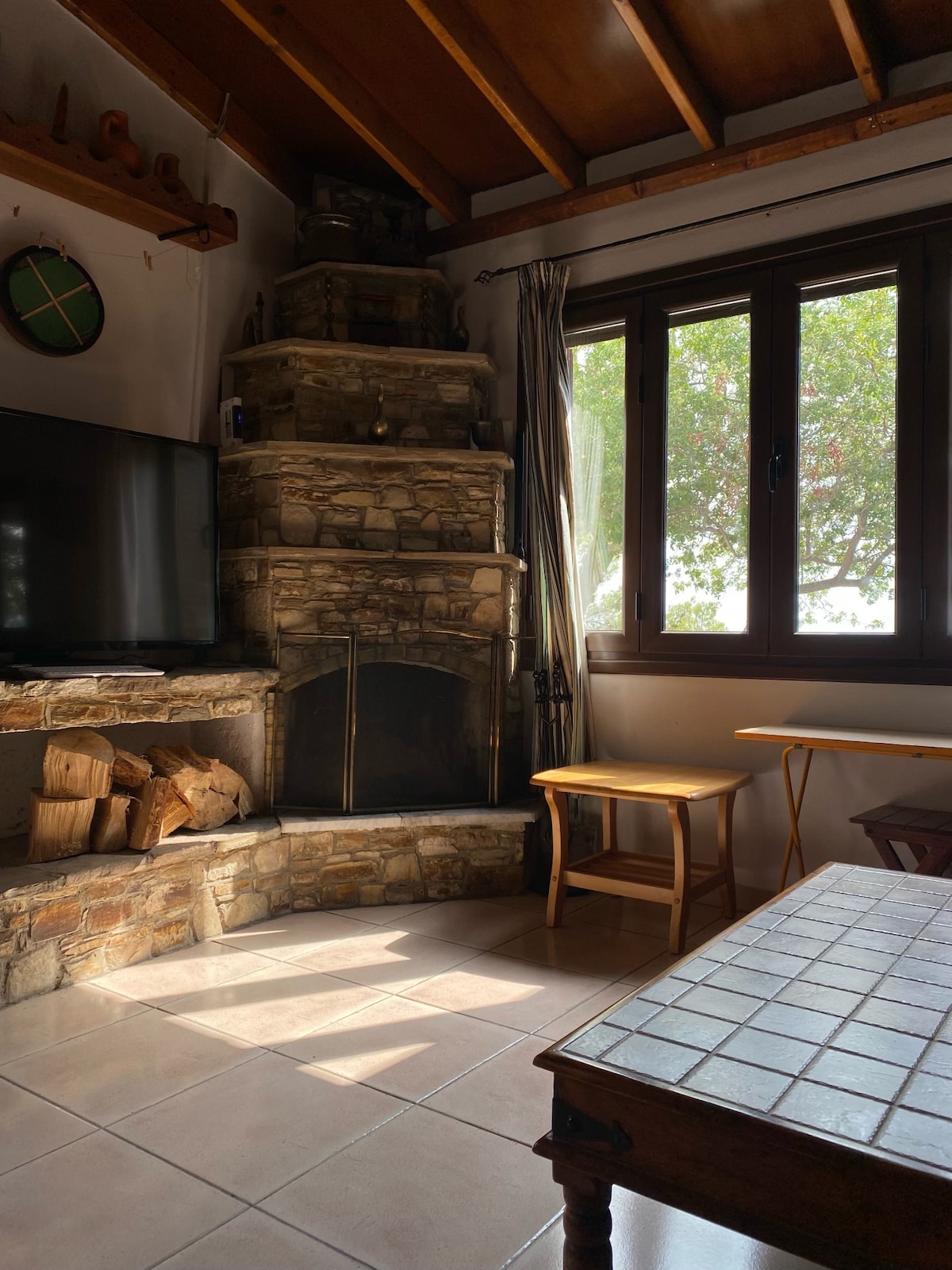 A stone fireplace with a rustic design is featured prominently, surrounded by a cozy seating area. Natural light streams through the wooden-framed window, illuminating the tiled floor. A small wooden table and additional seating are positioned beside the fireplace, creating a warm gathering space.