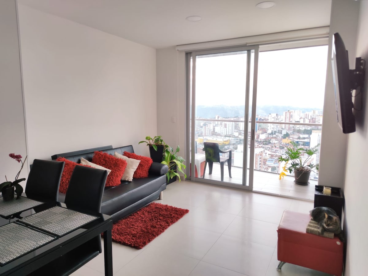 A spacious living area is furnished with a black sofa adorned with red cushions, complemented by a dining table and chairs. Large glass doors allow for ample natural light and reveal a mountain view. Indoor plants and a small rug add a touch of color.
