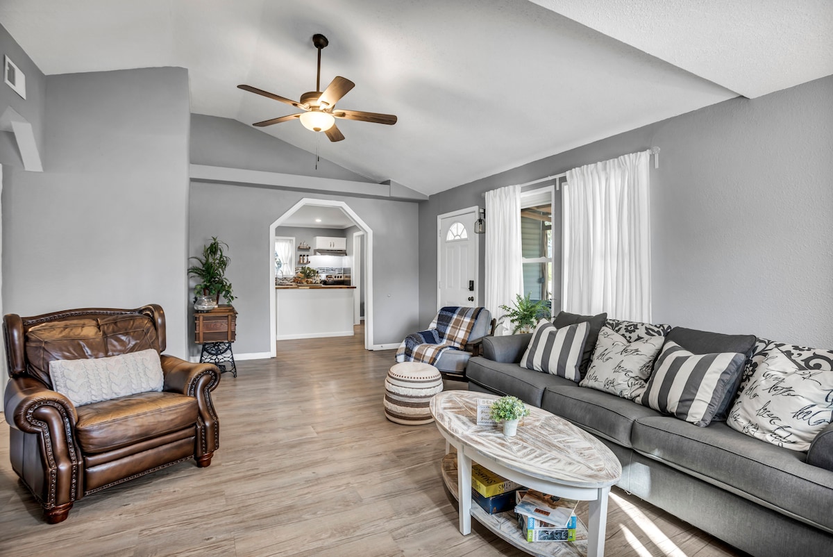 A well-lit living area features a mixture of comfortable seating options, including a leather armchair and a large sofa adorned with patterned cushions. A round coffee table is situated in the center, with a few books displayed. A ceiling fan adds a functional element to the space.