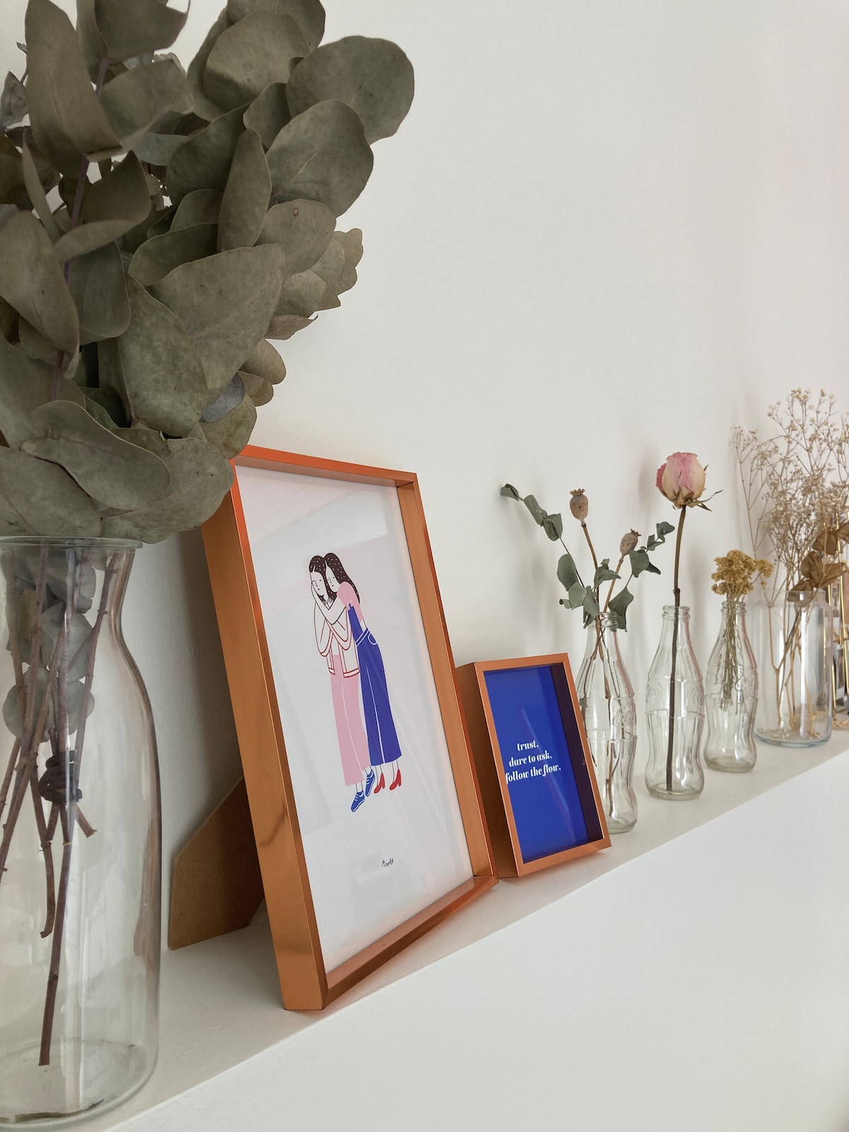 A shelf displays an arrangement of decorative items including a framed illustration, several slender glass vases containing dried flowers, and a small blue framed message. Eucalyptus branches in a tall vase add an earthy touch to the display.