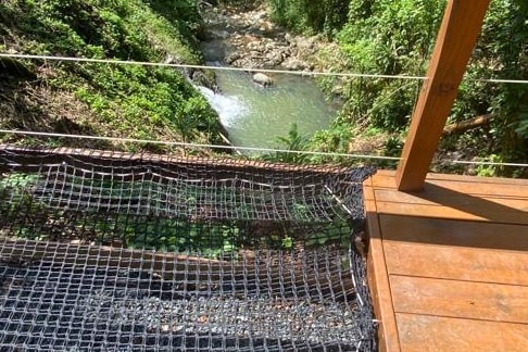 A wooden deck overlooks a lush green area with a flowing stream visible below. The natural surroundings feature dense foliage, creating a sense of seclusion. A safety net is positioned along the edge of the deck, providing security while enjoying the view.