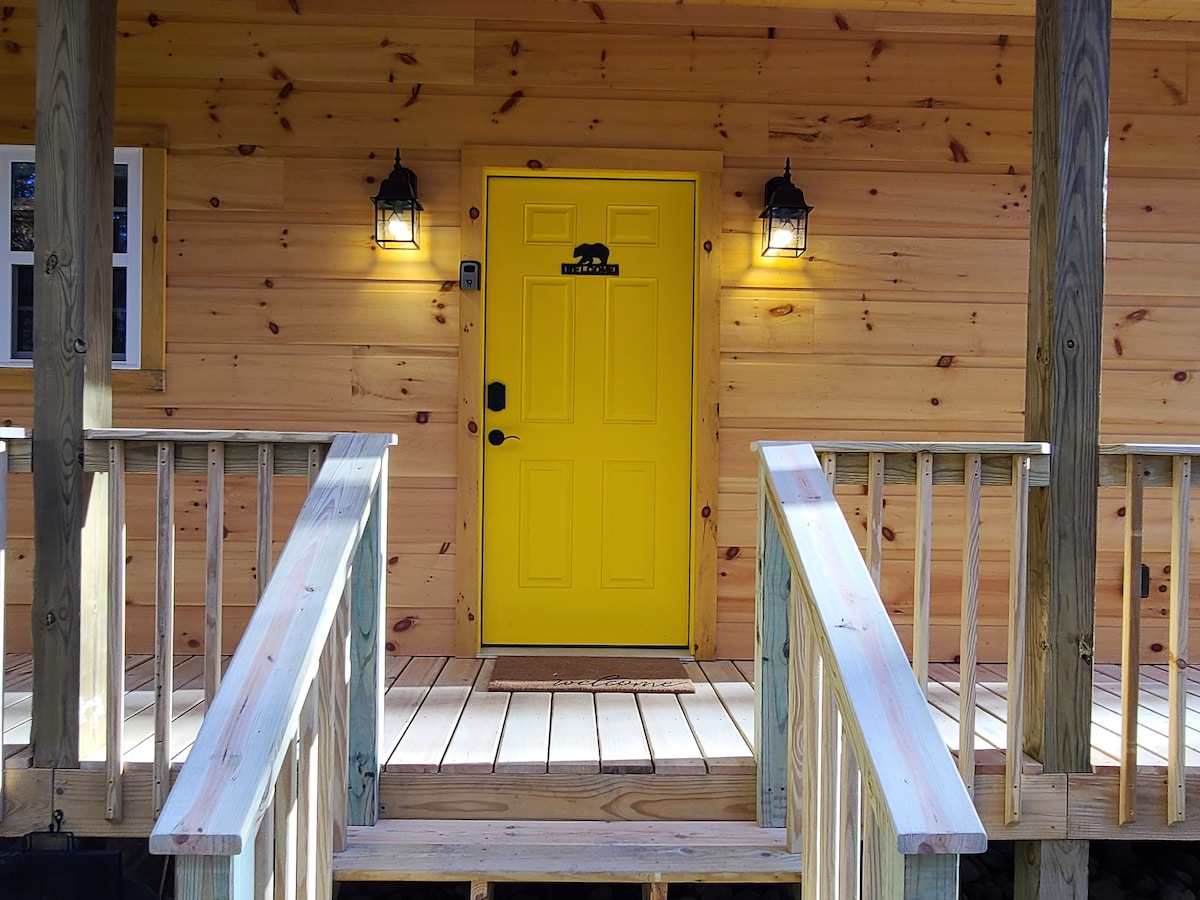 The entrance features a vibrant yellow door framed by warm wooden siding. Two lanterns provide illumination on either side, while a welcome mat is placed on the porch. The ramp leads to the front door, enhancing accessibility.