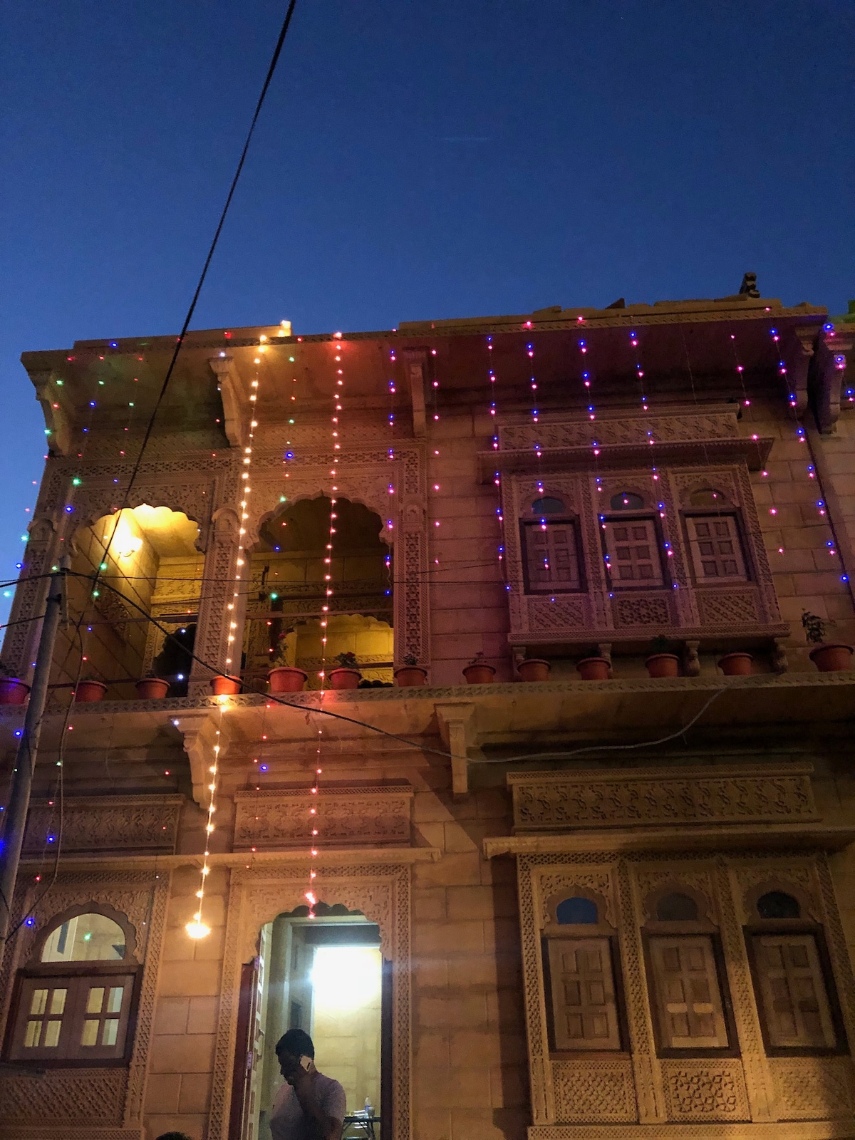 The three-story facade of a boutique hotel is showcased, featuring intricately carved golden stone. Colorful lights hang from the balcony and windows, enhancing the festive atmosphere. Potted plants are displayed along the ledges, adding greenery to the charming exterior.