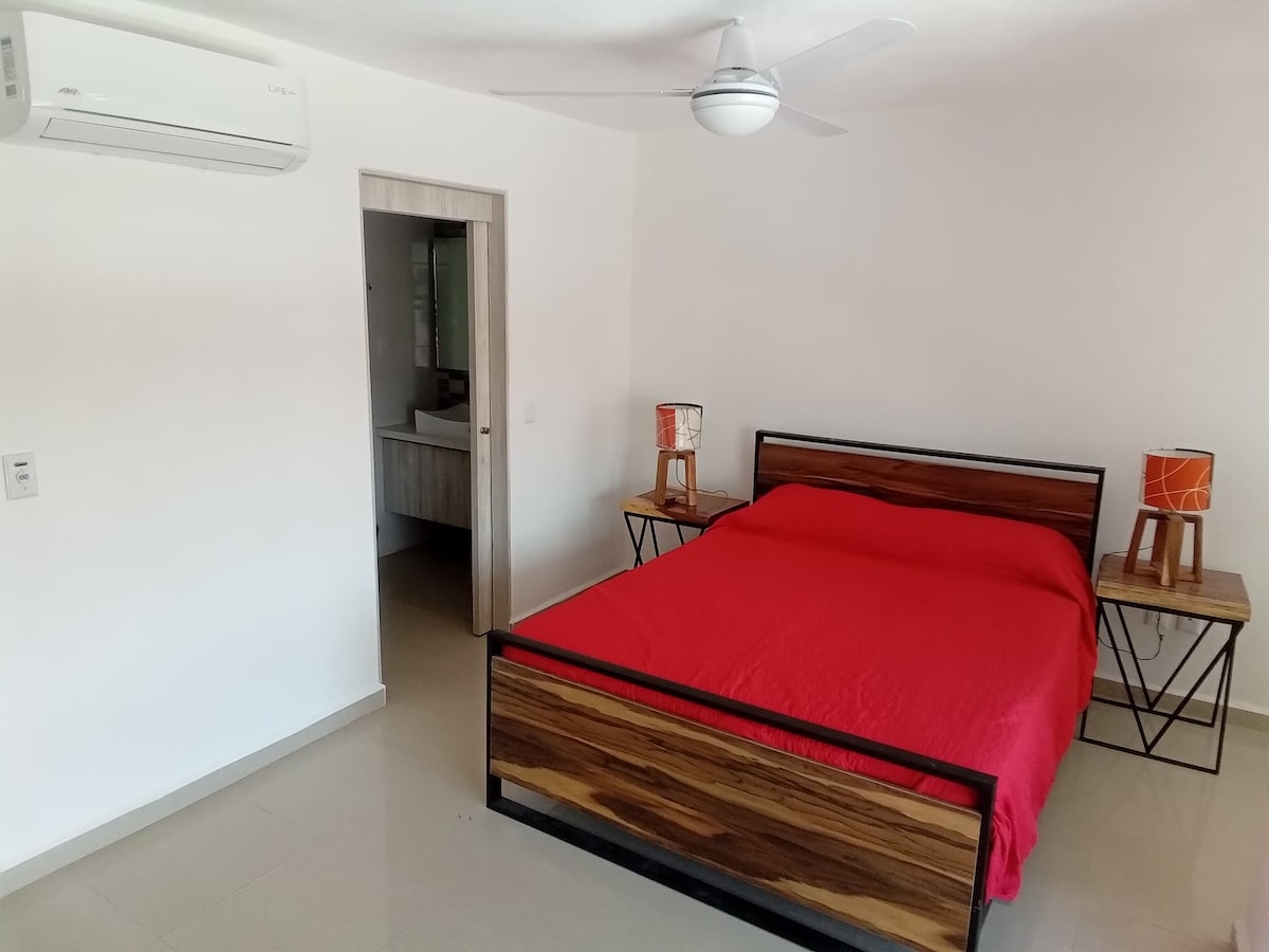 A bright bedroom is furnished with a spacious bed dressed in a vibrant red coverlet. Two nightstands with unique lamps sit on either side, and a ceiling fan is mounted above. A doorway leads to a bathroom area, enhancing the room's functionality.