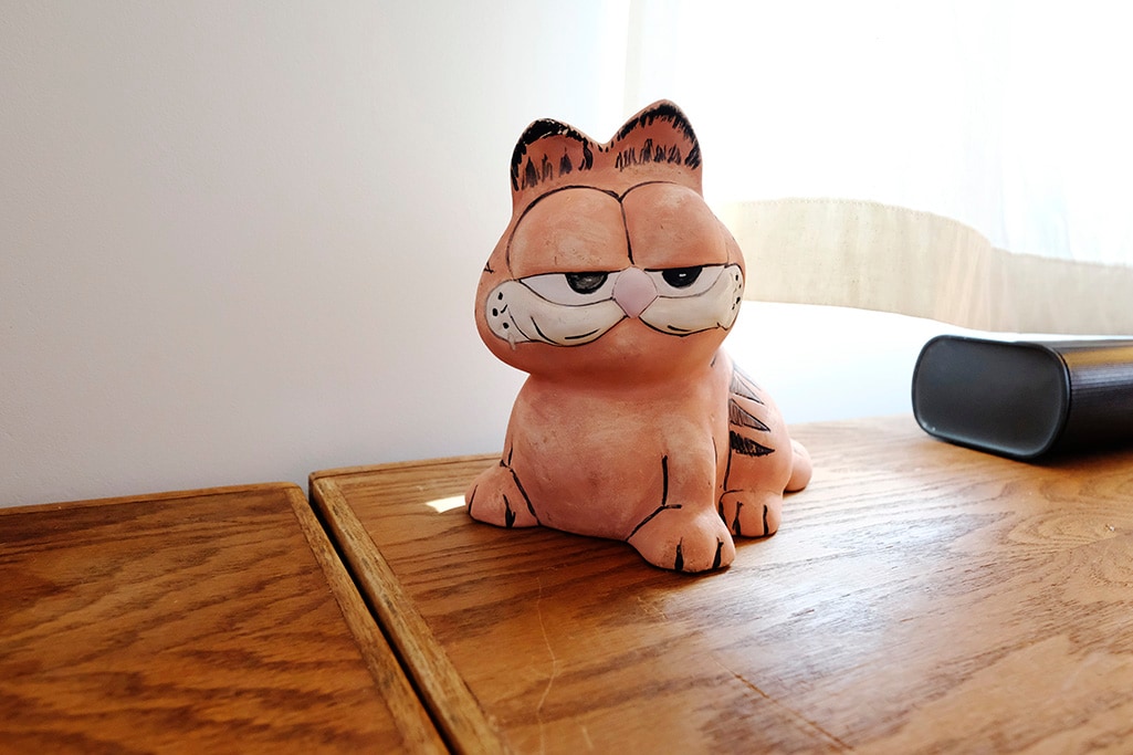 A ceramic figurine of a cat, resembling a well-known cartoon character, is displayed on a wooden surface. The cat features a slightly smug expression, with detailing on its fur and facial features. Natural light filters in from a nearby window, illuminating the scene.