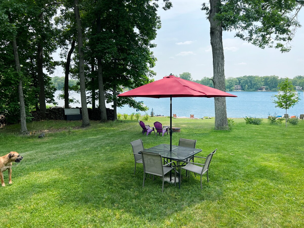 In the foreground, a patio dining set is situated under a large umbrella on the expansive grass yard. To the side, a dog is seen gazing towards the tranquil lake, which is framed by trees and features colorful chairs along the waterfront.