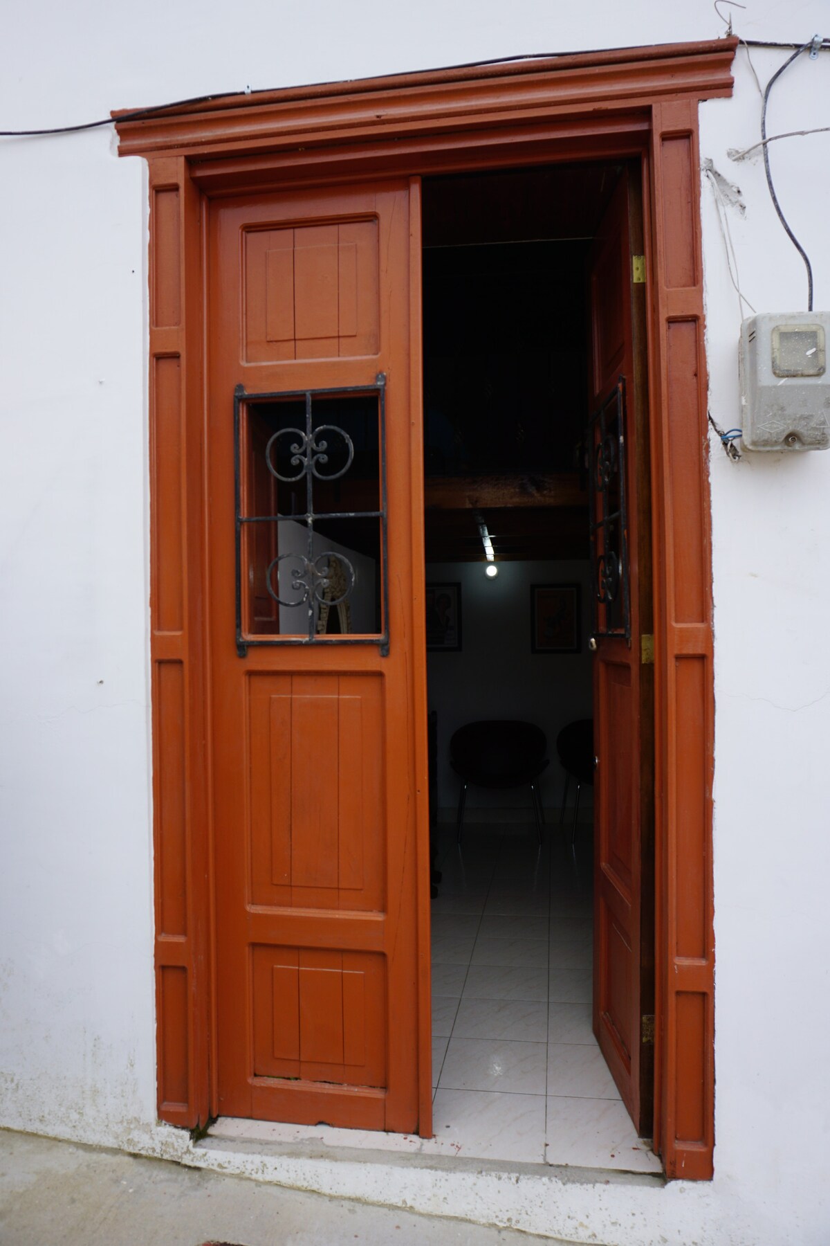 A wooden door with intricate ironwork stands ajar, revealing a glimpse of the interior. The door's warm orange hue contrasts with the white exterior wall, while a tiled floor is visible inside, contributing to a welcoming entrance.