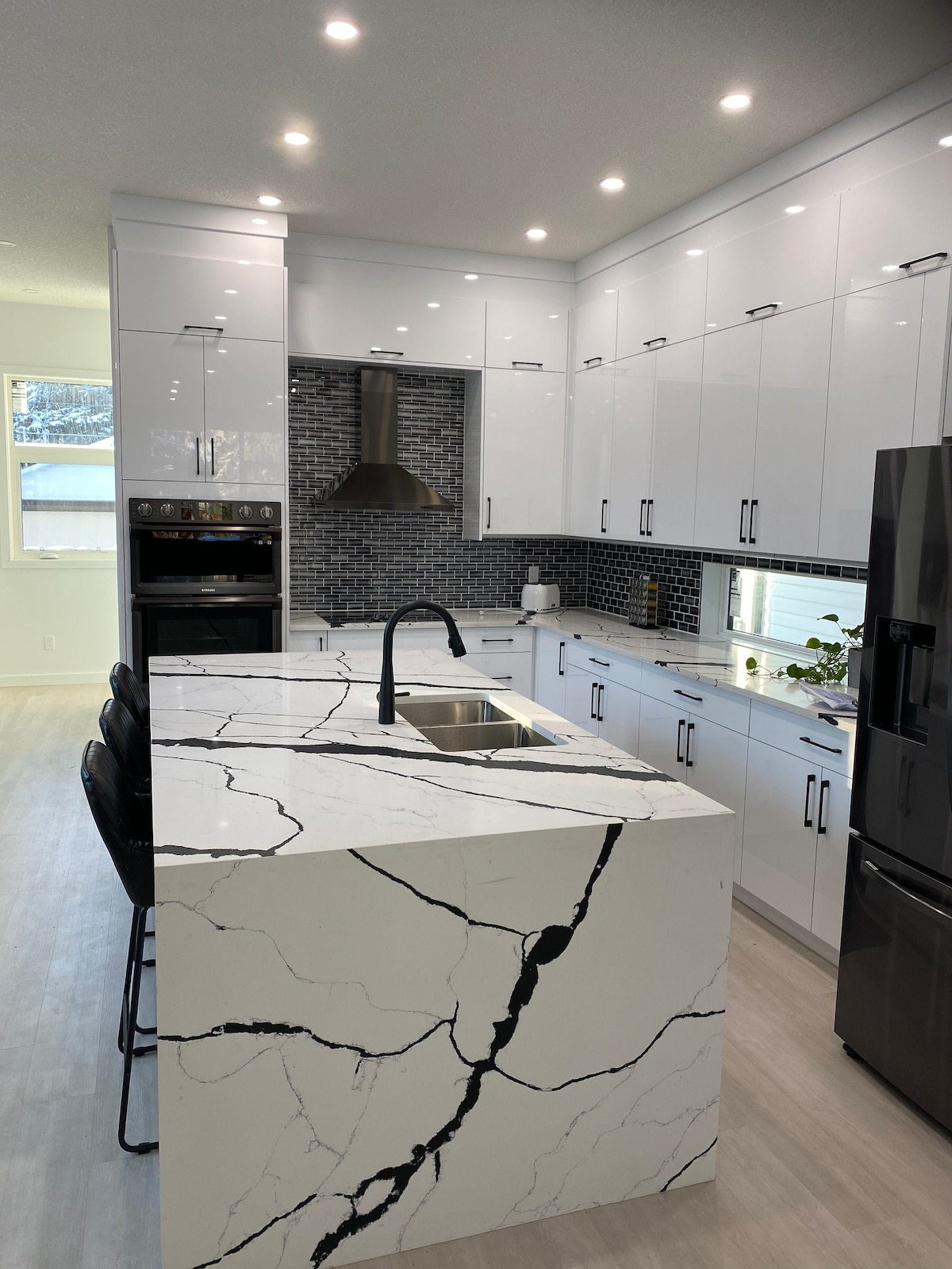 A modern kitchen showcases sleek white cabinetry and a striking countertop featuring bold black veins. A spacious island with seating is positioned in the center, highlighted by under-cabinet lighting and a stylish range hood above the stove. Natural light filters through nearby windows.