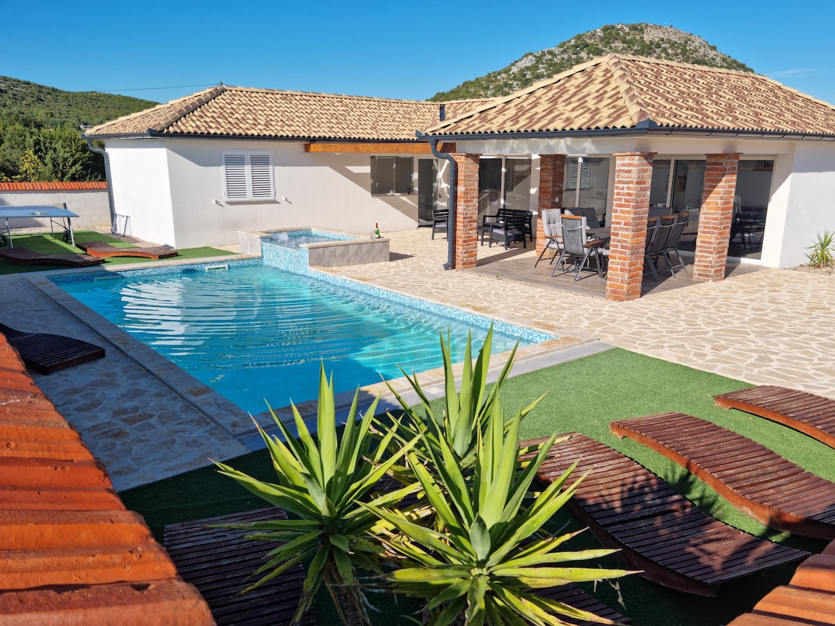 A tranquil outdoor area features a heated swimming pool surrounded by lounge chairs and greenery. A covered patio is visible, furnished with a dining table and chairs, providing a space for outdoor meals. The landscape includes mountains in the background.