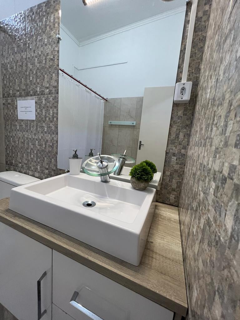 A modern bathroom features a minimalist sink with a square basin set on a wooden countertop. Elegant mosaic tiles cover the walls, while a small decorative plant adds a touch of greenery. Natural light illuminates the space, enhancing its clean lines and fresh feel.
