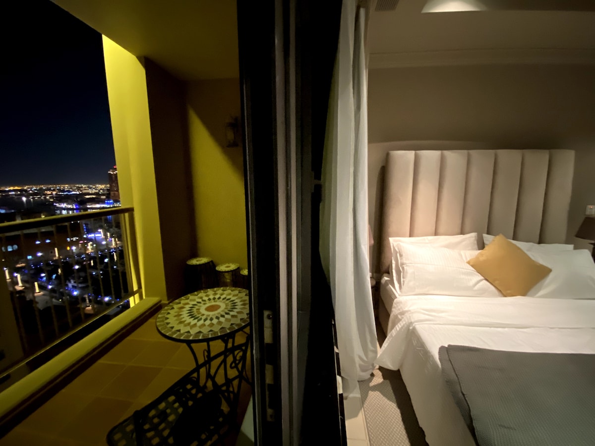 A cozy bedroom presents a spacious bed with light-colored linens and a decorative accent pillow. Adjacent, a balcony area features a round table and two stools, offering an outdoor seating option with a view of the city lights at night.