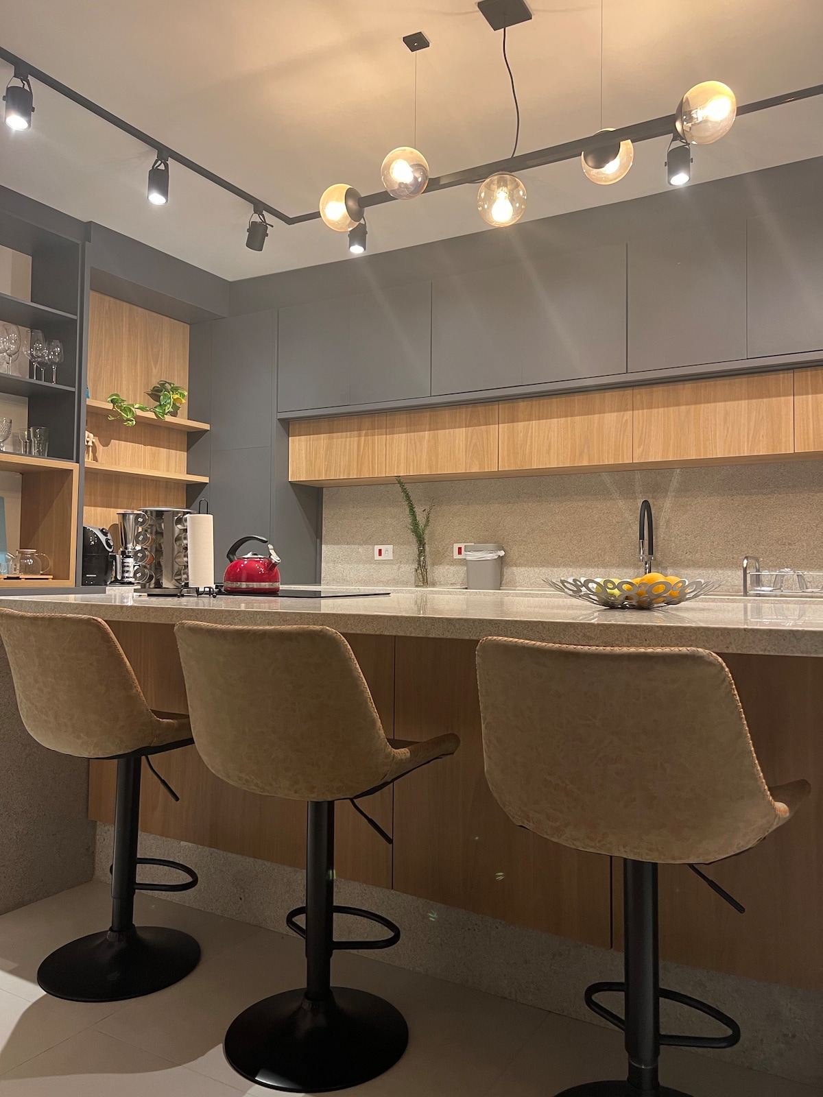 A modern kitchen showcases a mix of wooden and gray cabinetry, providing ample storage. Two high-back stools are arranged at a sleek countertop. Task lighting highlights the space, while an elegant fruit bowl sits center stage. Appliances, including a coffee maker, are neatly arranged.