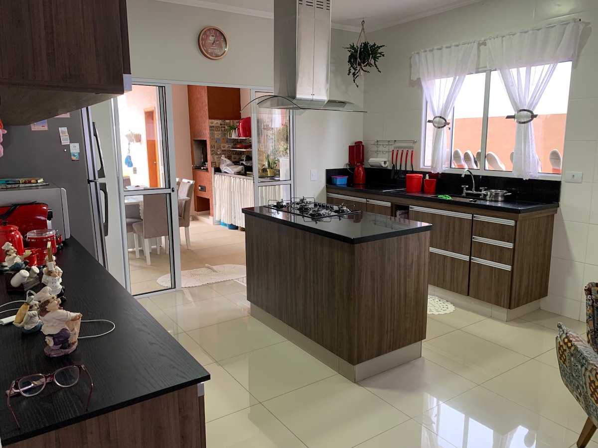 A modern kitchen is featured with wooden cabinetry and a large island in the center. Stainless steel appliances, including a refrigerator and oven, complement the sleek design. Natural light floods the space through two large windows, enhancing the open layout.