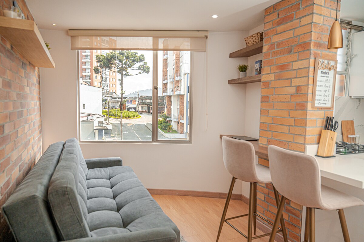 A spacious lounge area features a comfortable green sofa, positioned against a textured brick wall. Natural light filters through a large window, offering a view of the outdoor surroundings. A modern kitchen bar with stools complements the space, providing additional seating.