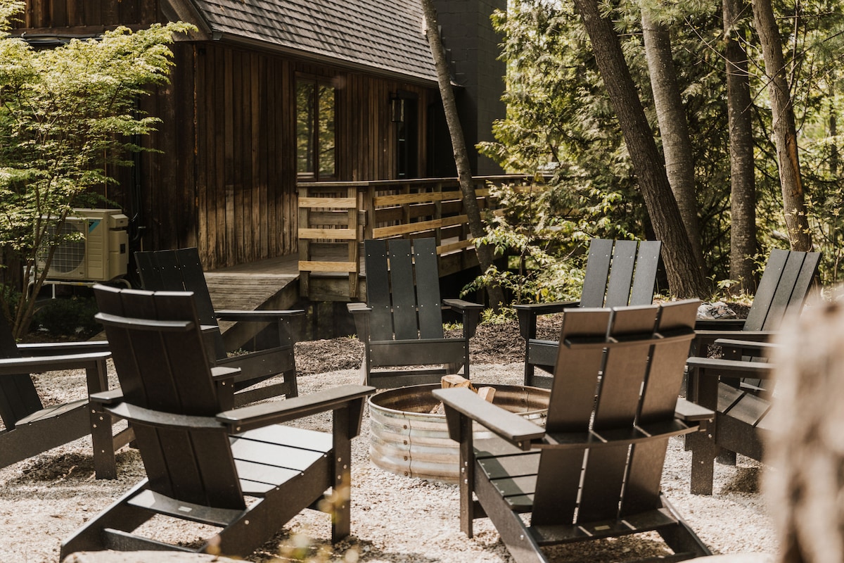 A fire pit is surrounded by several dark wooden Adirondack chairs, inviting warmth and conversation. The rustic exterior of the barn house is visible in the background, alongside lush greenery and a wooden deck that adds to the outdoor ambiance.