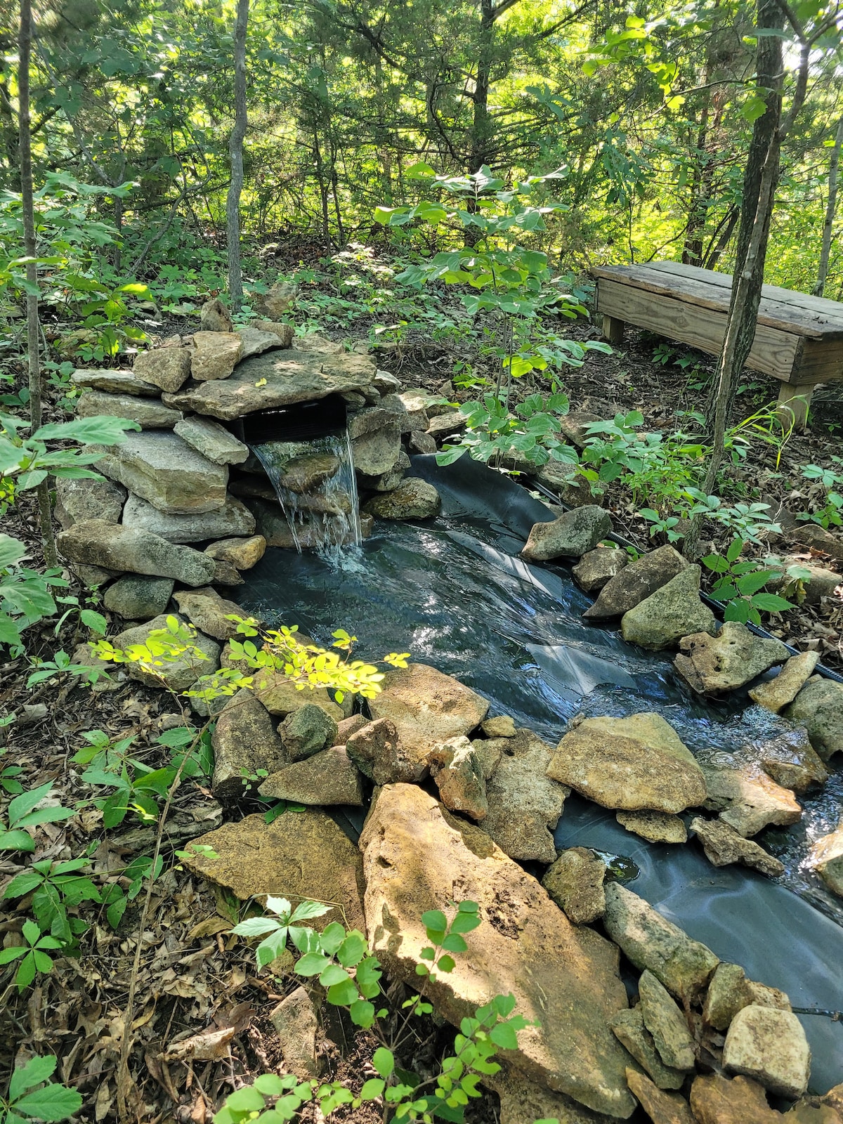 A serene waterfall feature is surrounded by natural stone, with water flowing over the rocks into a small stream. Lush greenery and trees frame the scene, creating a calming atmosphere in the wooded area.