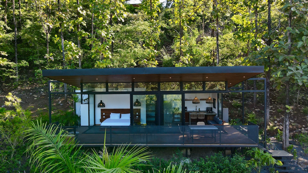 A modern guest house is surrounded by lush greenery, featuring large glass walls that provide views of the natural landscape. A spacious deck extends in front, while sleek hanging lights and wooden accents contribute to the contemporary design.
