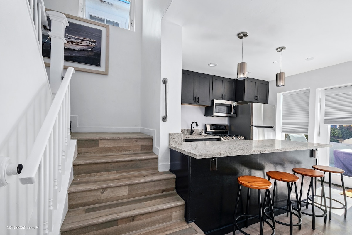 A modern kitchen space features sleek black cabinetry and stainless steel appliances. A white countertop offers seating for four on wooden stools. Natural light enters through nearby windows, illuminating the open-concept layout. A staircase leads to the upper floor, enhancing the airy atmosphere.