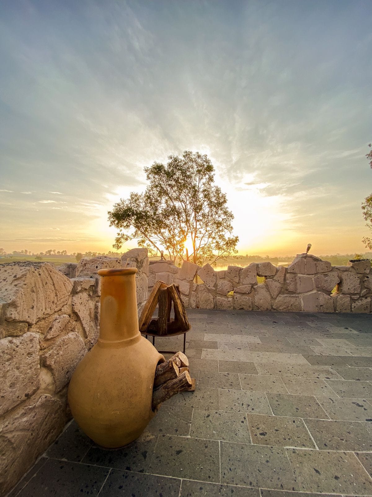 A large clay pot is positioned next to a stack of firewood on a stone patio. In the background, a tree silhouettes against a soft sunrise, with the light creating an inviting ambiance across the landscape.