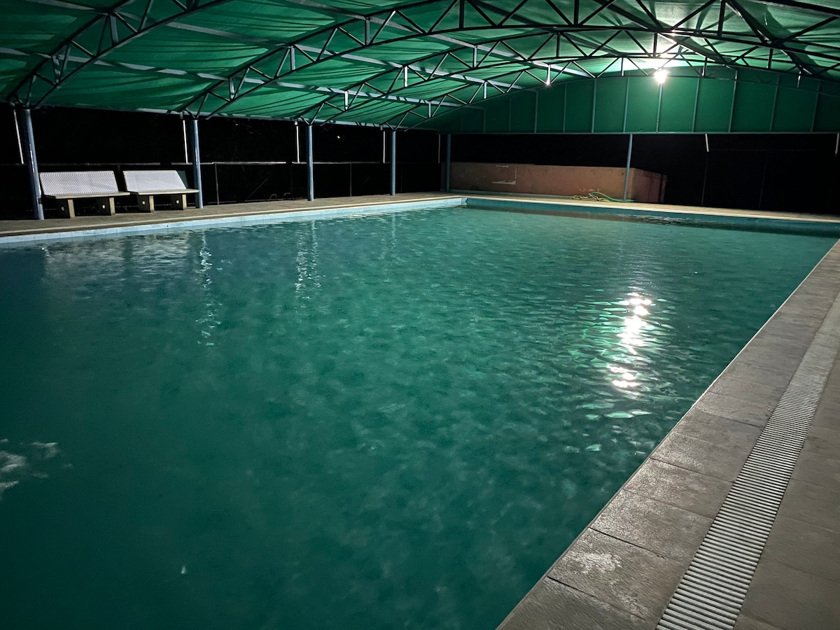 An outdoor swimming pool is showcased under a covered area, illuminated by overhead lights. The water reflects various shades of blue and green, with a few lounge chairs positioned nearby. The serene environment is enhanced by the surrounding darkness.