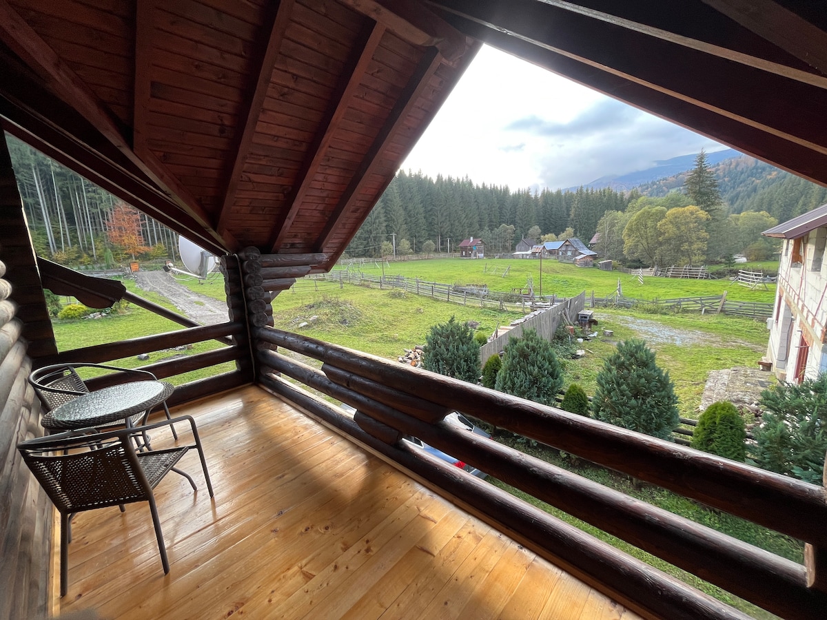 A covered balcony features wooden beams and a rustic floor, offering a small round table and two chairs. The view includes a lush, green field bordered by trees and distant cabins, creating a picturesque natural setting.