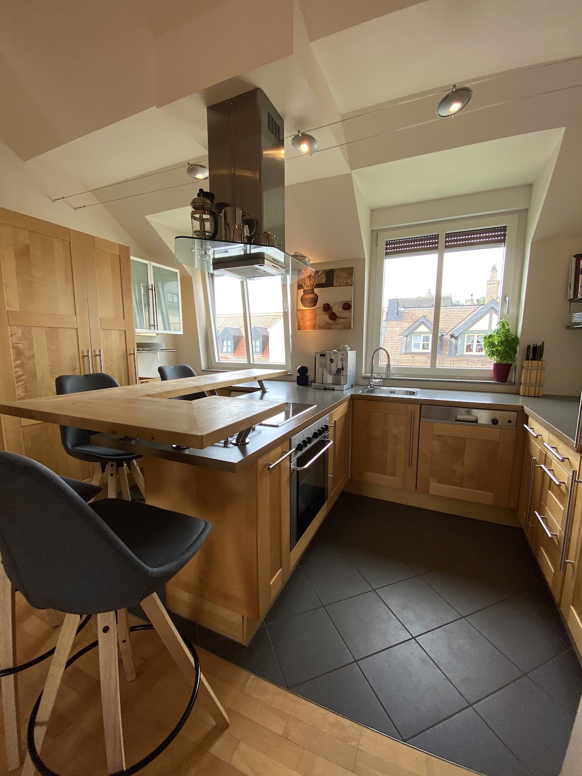 A modern kitchen with wooden cabinetry features an island with bar stools. Equipped with stainless steel appliances, the area is illuminated by natural light through a window. The sleek countertop offers ample space for meal preparation, complemented by a decorative plant on the windowsill.