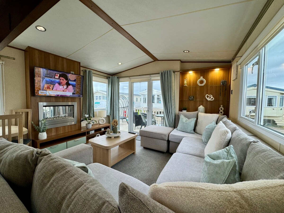 A welcoming living area features a spacious sectional sofa with light-colored cushions and a coffee table at the center. A television is mounted on the wall, and large windows provide natural light. Decor elements are displayed on a feature wall, enhancing the cozy atmosphere.