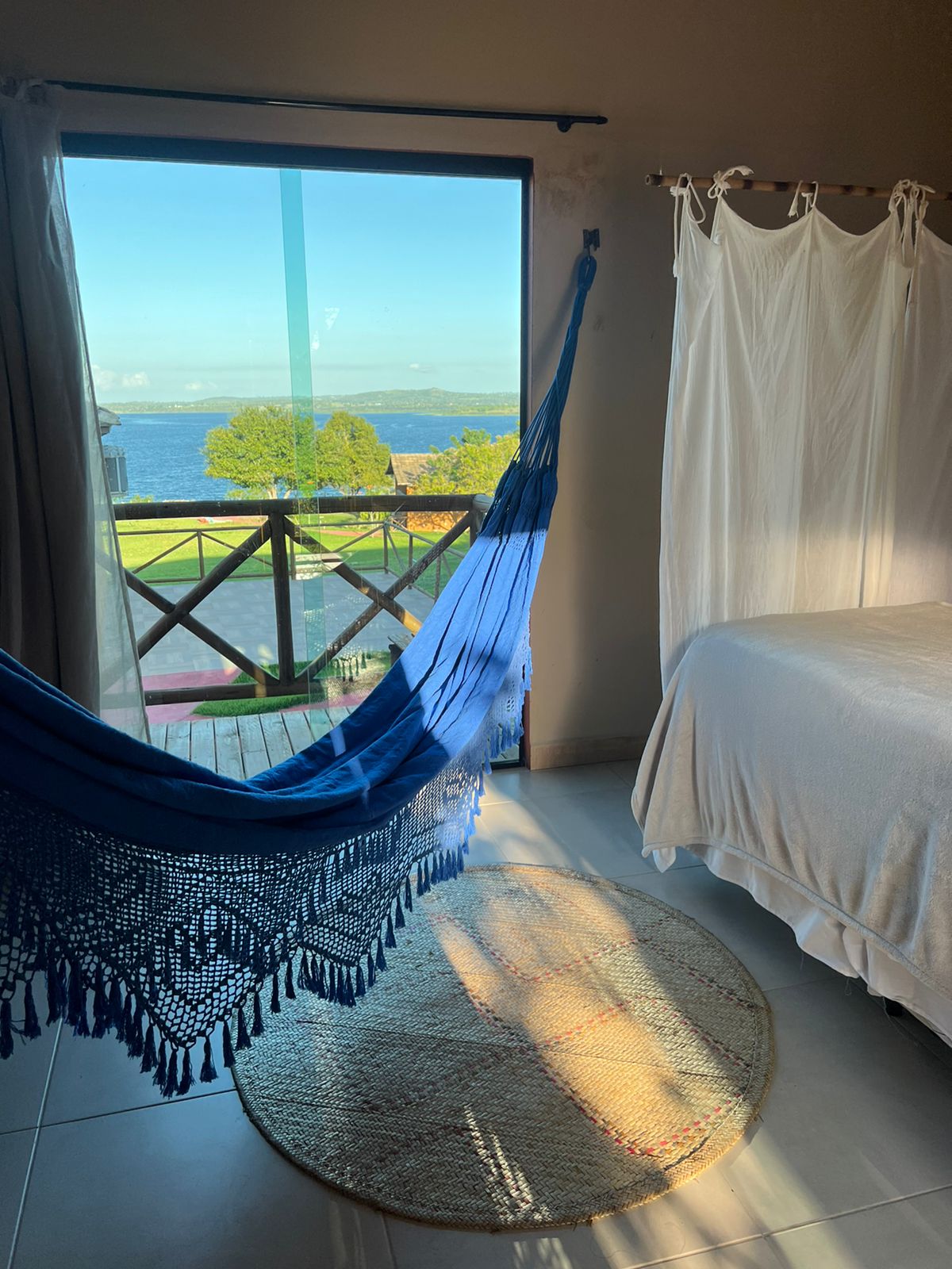 A blue hammock hangs beside a large window, revealing views of the river and landscape beyond. Sunlight streams into the room, casting gentle patterns onto a circular woven rug placed on the tiled floor, next to a spacious bed dressed in white linens.