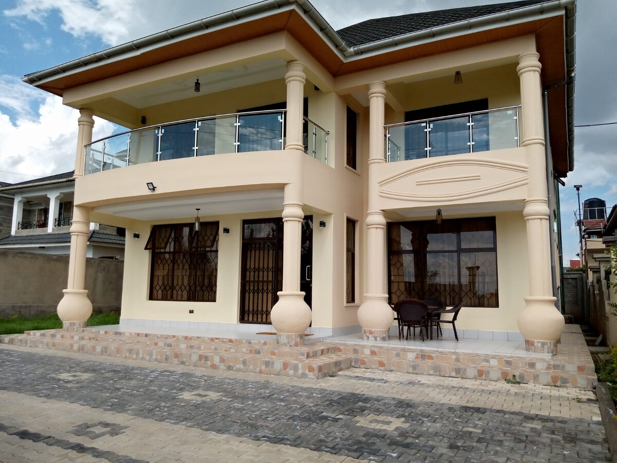 A two-story house with a cream-colored exterior is showcased. Balconies with glass railings line the front, complemented by decorative pillars. A seating area with a table and chairs is visible on the ground floor, and the entrance is framed by a spacious porch.
