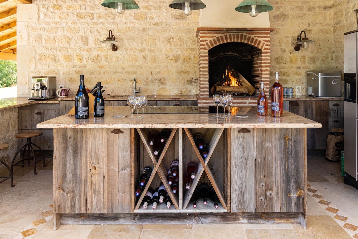 An outdoor kitchen features a large wooden island with a marble countertop, displaying several bottles of wine. An integrated wine rack holds multiple bottles underneath. A fireplace is set into the stone wall, showcasing an inviting fire, while overhead lights provide illumination.