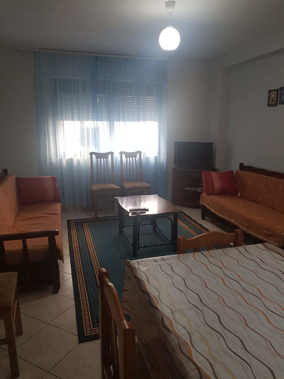 A living area is furnished with two armchairs and two sofas, arranged around a central coffee table. A patterned area rug lies on the tiled floor. Natural light enters through a window, which is covered with light curtains. A television is positioned in one corner.