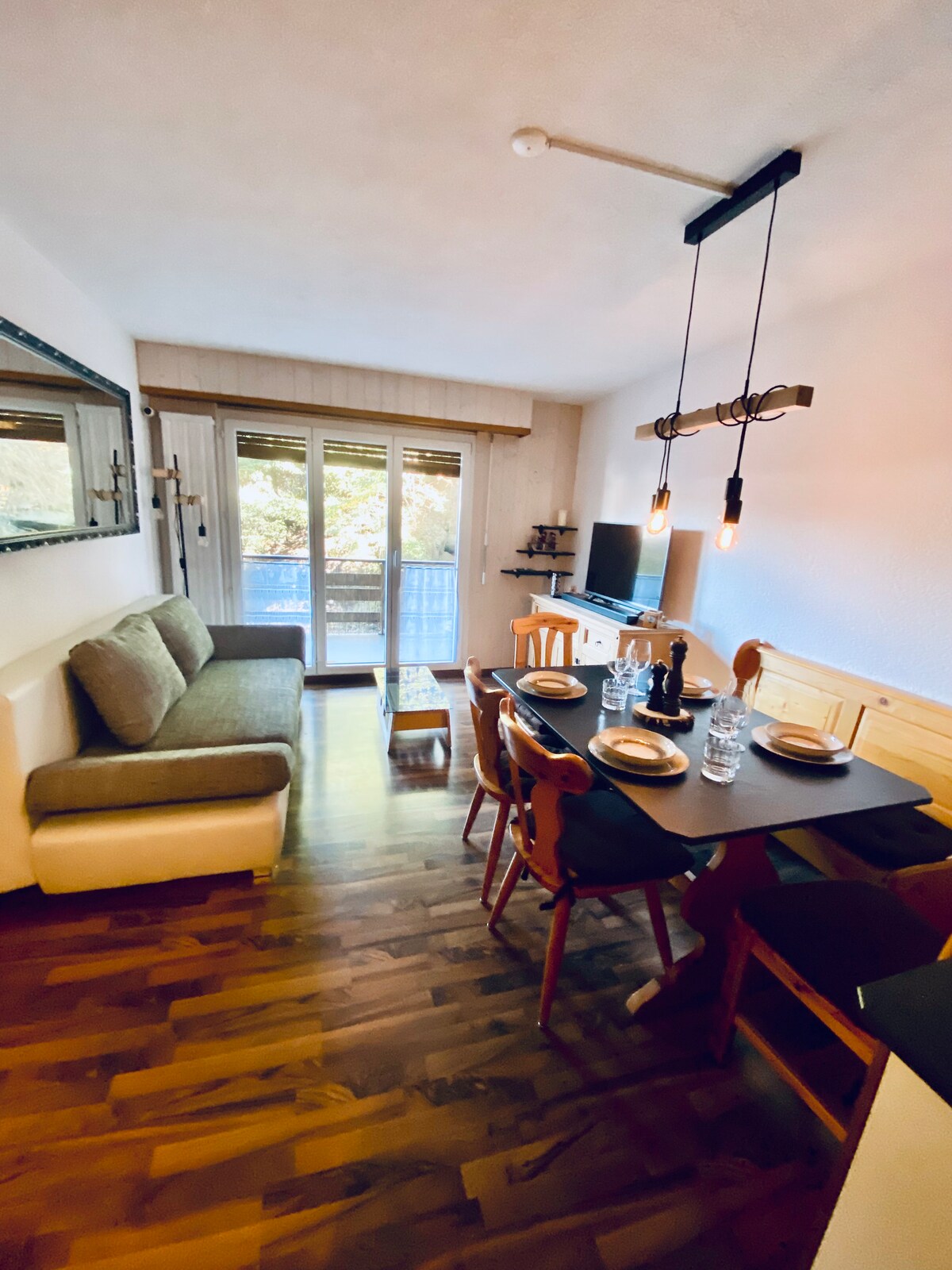 A cozy living area is visible, featuring a comfortable sofa near a set of glass doors leading to a balcony. A dining table with chairs is set for a meal, complemented by an elegant light fixture above. Natural light illuminates the warm wooden flooring.