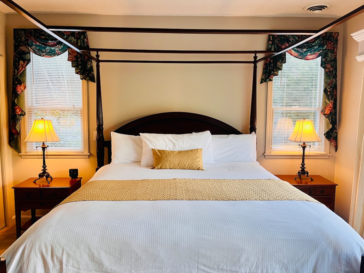A king-sized bed is positioned centrally in the room, complemented by two bedside tables with lamps. Natural light enters through two windows adorned with floral curtains, creating a soothing ambience. A decorative golden accent pillow is placed on the bed, enhancing the overall elegance.