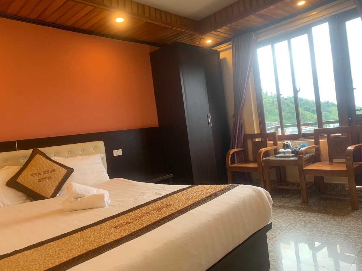 A well-appointed bedroom is presented, featuring a spacious bed with a decorative throw and two wooden chairs by a window. Natural light fills the room, which is accented by a warm orange wall and light wooden ceiling.