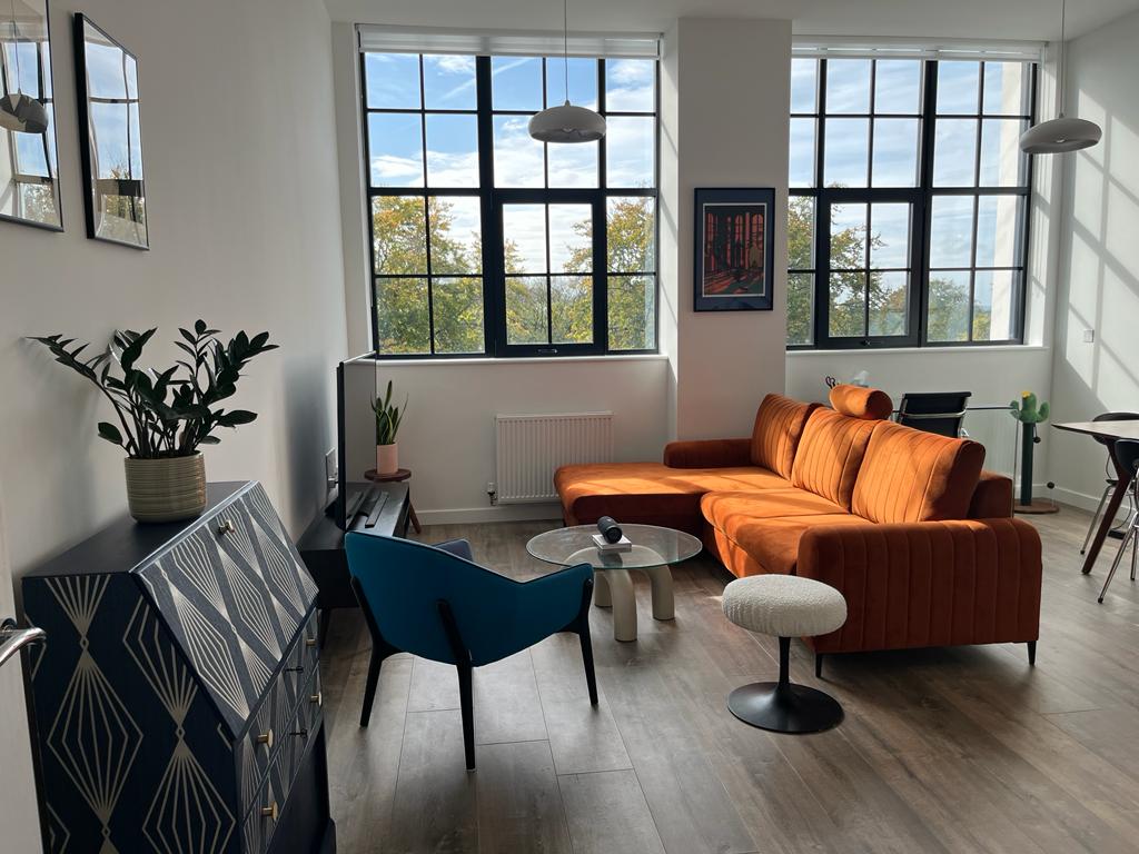 A modern living area features large industrial windows that allow natural light to fill the room. An orange sectional sofa invites relaxation, accompanied by a round coffee table and a blue accent chair. A flat-screen TV is positioned on a stylish cabinet, completing the space.