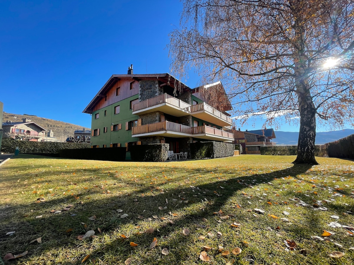 A charming three-story structure is set against a clear blue sky, surrounded by a lush green lawn adorned with fallen leaves. Balconies are visible on each level, enhancing the building's inviting exterior. The peaceful landscape suggests a tranquil environment for relaxation.