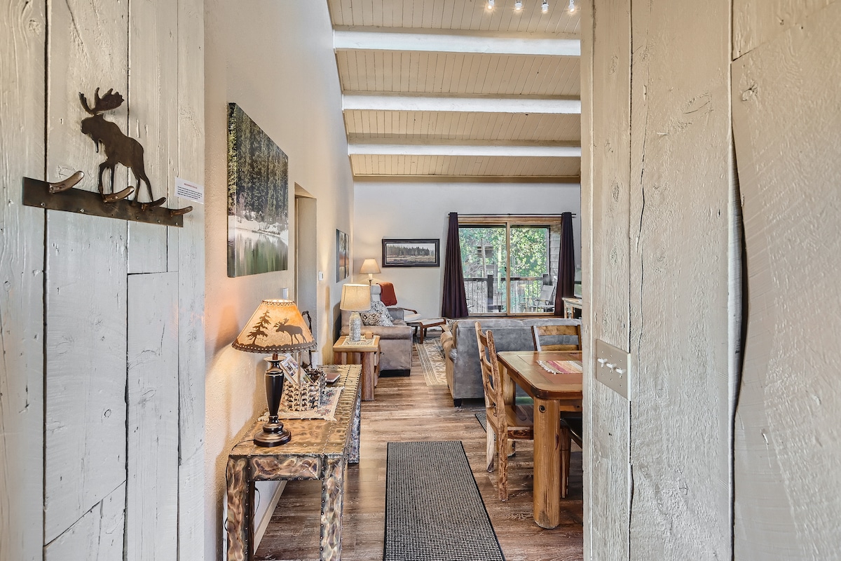 A spacious living area is observed with vaulted ceilings and wooden beams. Natural light floods through large windows, illuminating rustic furnishings and a cozy seating arrangement. A dining table crafted from natural wood complements the design, alongside lamps that provide ambient lighting.