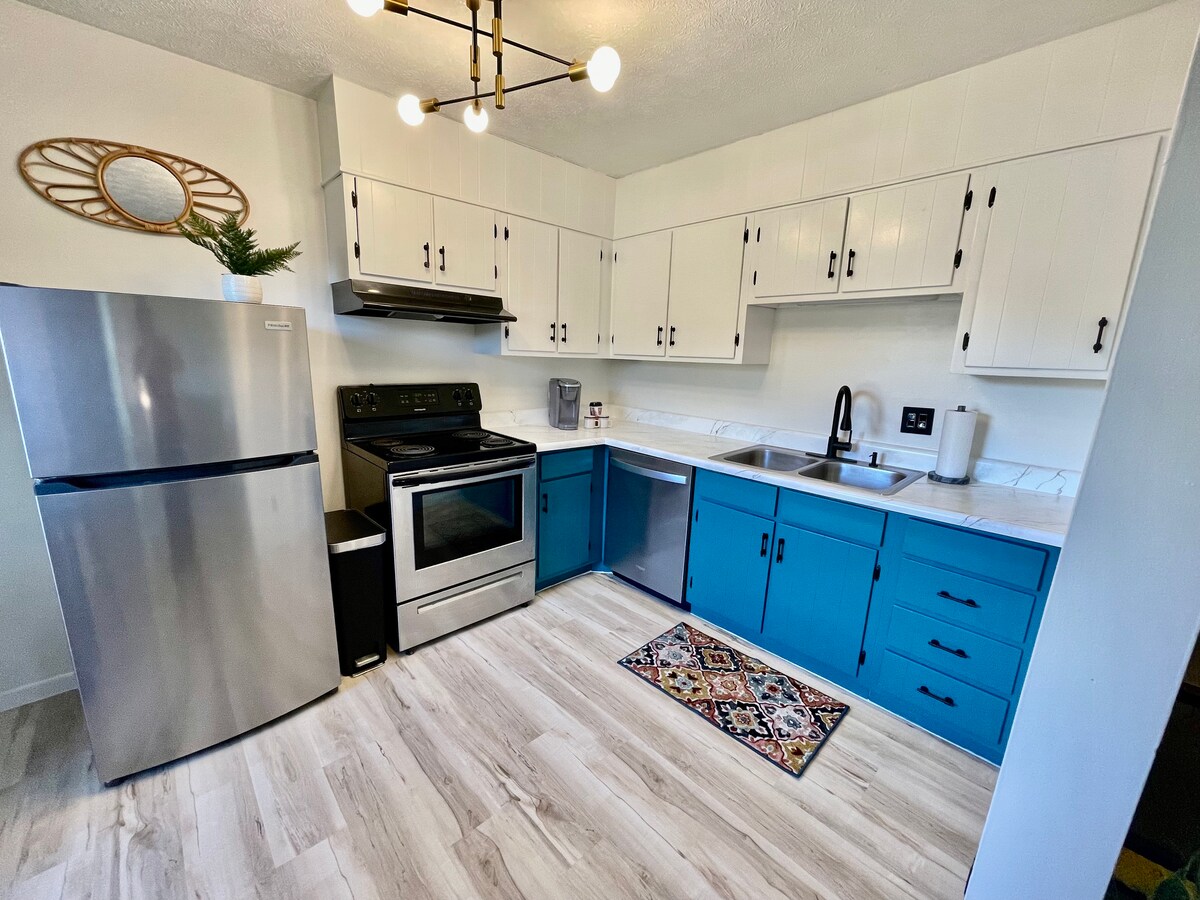 A modern kitchen is highlighted by blue lower cabinets and white upper cabinets, providing a fresh contrast. Stainless steel appliances, including a refrigerator and oven, add functionality. A patterned rug is placed in front of the sink, enhancing the workspace within the bright, airy layout.