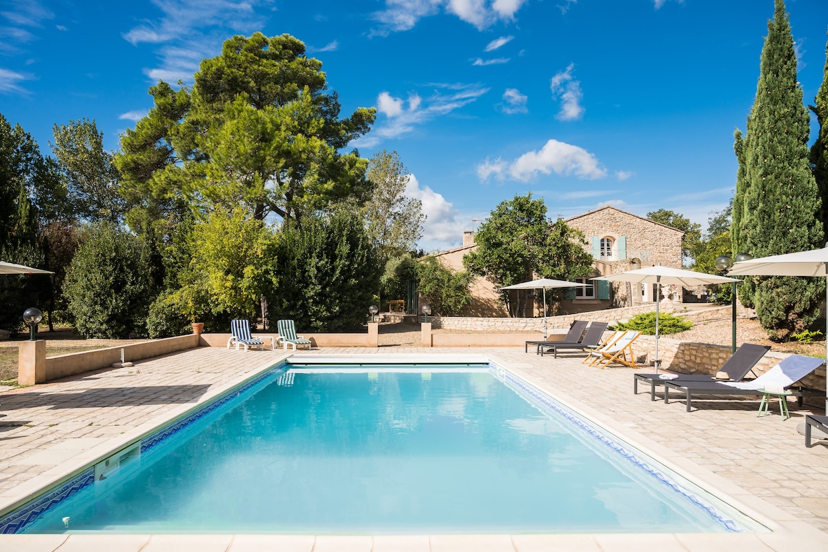 A clear blue swimming pool is surrounded by a stone patio, featuring several lounge chairs and umbrellas for shade. Lush greenery and trees provide a natural backdrop, while a large two-story building is visible in the distance.