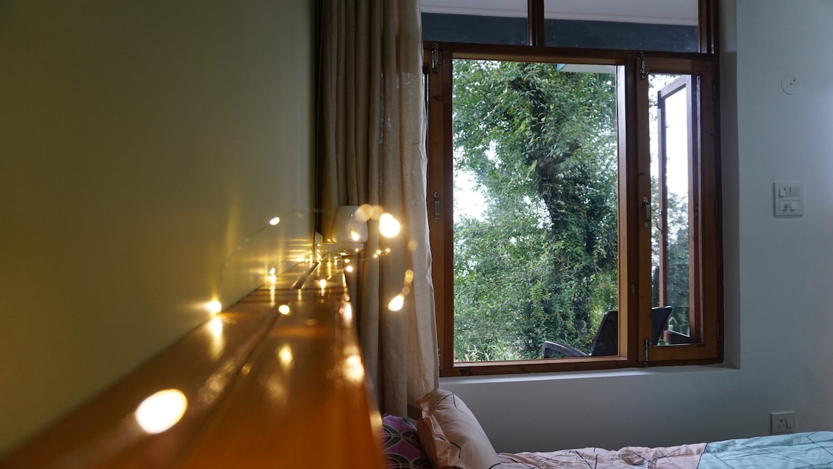 A cozy bedroom is framed by large windows, offering views of lush trees outside. Soft lighting from fairy lights is visible along the wooden headboard, creating a gentle glow. The room is designed to blend comfort with natural surroundings.