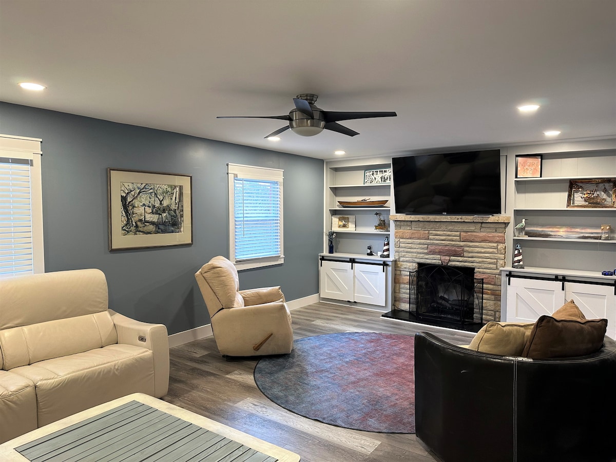 The spacious living area features a cozy seating arrangement with a cream-colored sofa and an ergonomic armchair positioned around a circular rug. A stone fireplace serves as a focal point, while built-in shelves flank the TV, providing a functional and stylish entertainment space.